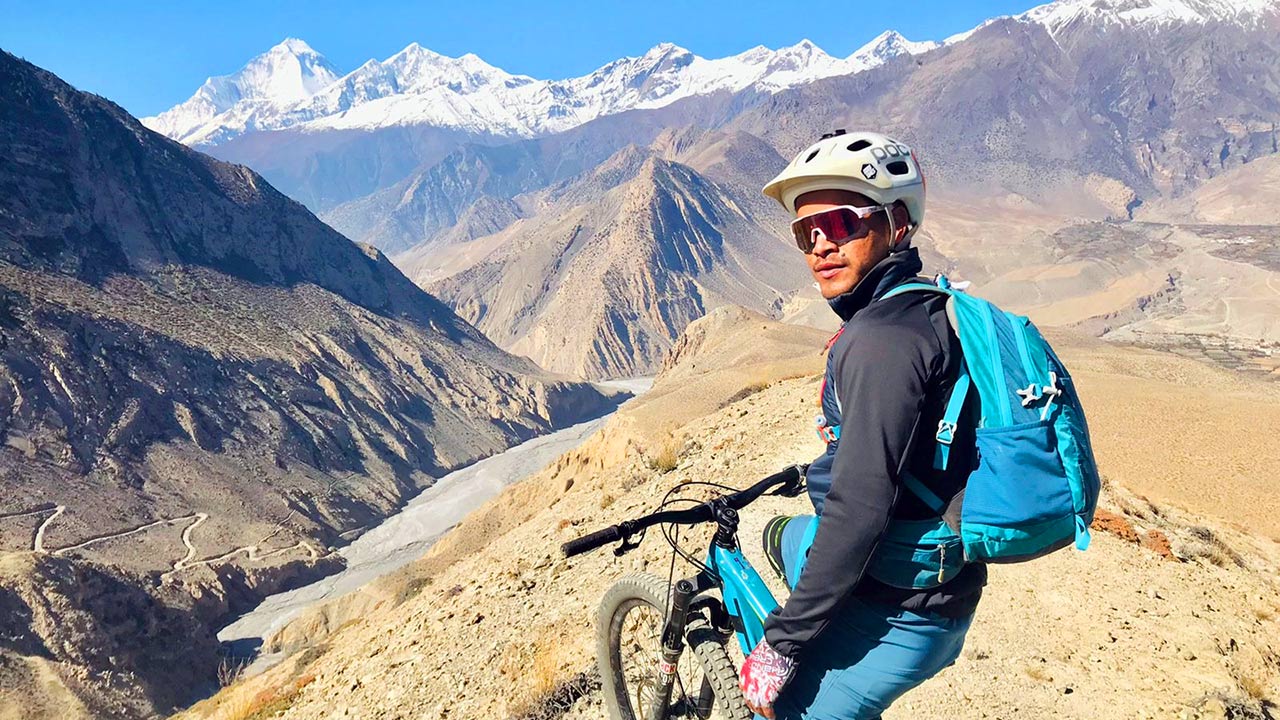 A mountain biker carrying a blue bag on his mountain bike poses for a picture at the Lupra single track with the view of Dhaulagiri and Tukuche peak in Mustang.