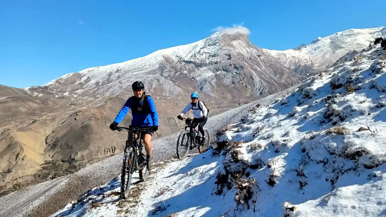 Two mountain bikers, the front with blue jacket and the rear one with the white jacket are descending down the Lupra single track covered with snow in Mustang. 