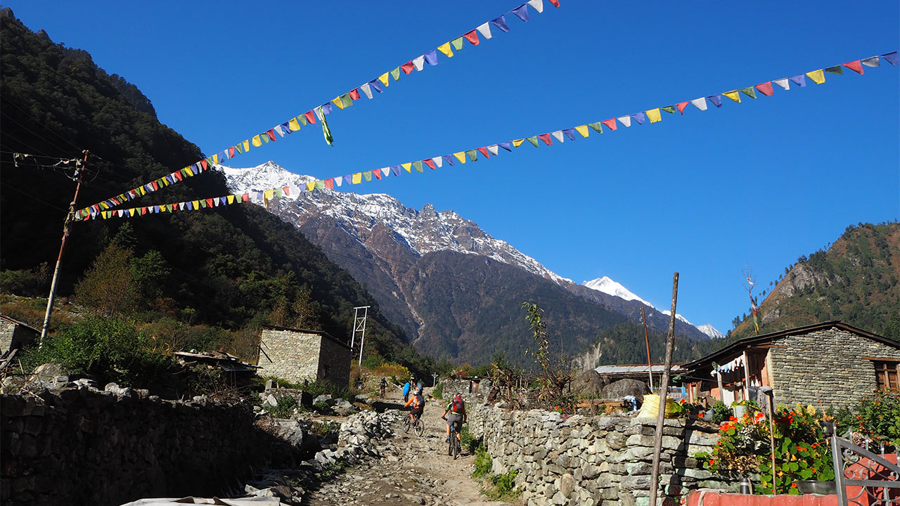 Two mountain bikers are feeling warmly welcomed with the Tibetan prayer flags hung in the air and the beautiful view of the mountains at Danaque in the Annapurna Circuit MTB tour in Nepal. 