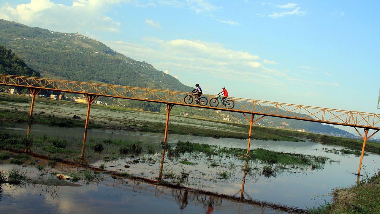 Two E-Mtb riders with blue-white jersey in the front and orange colored jersey on the back crossing over the yellow bride in Pame, Pokhara.