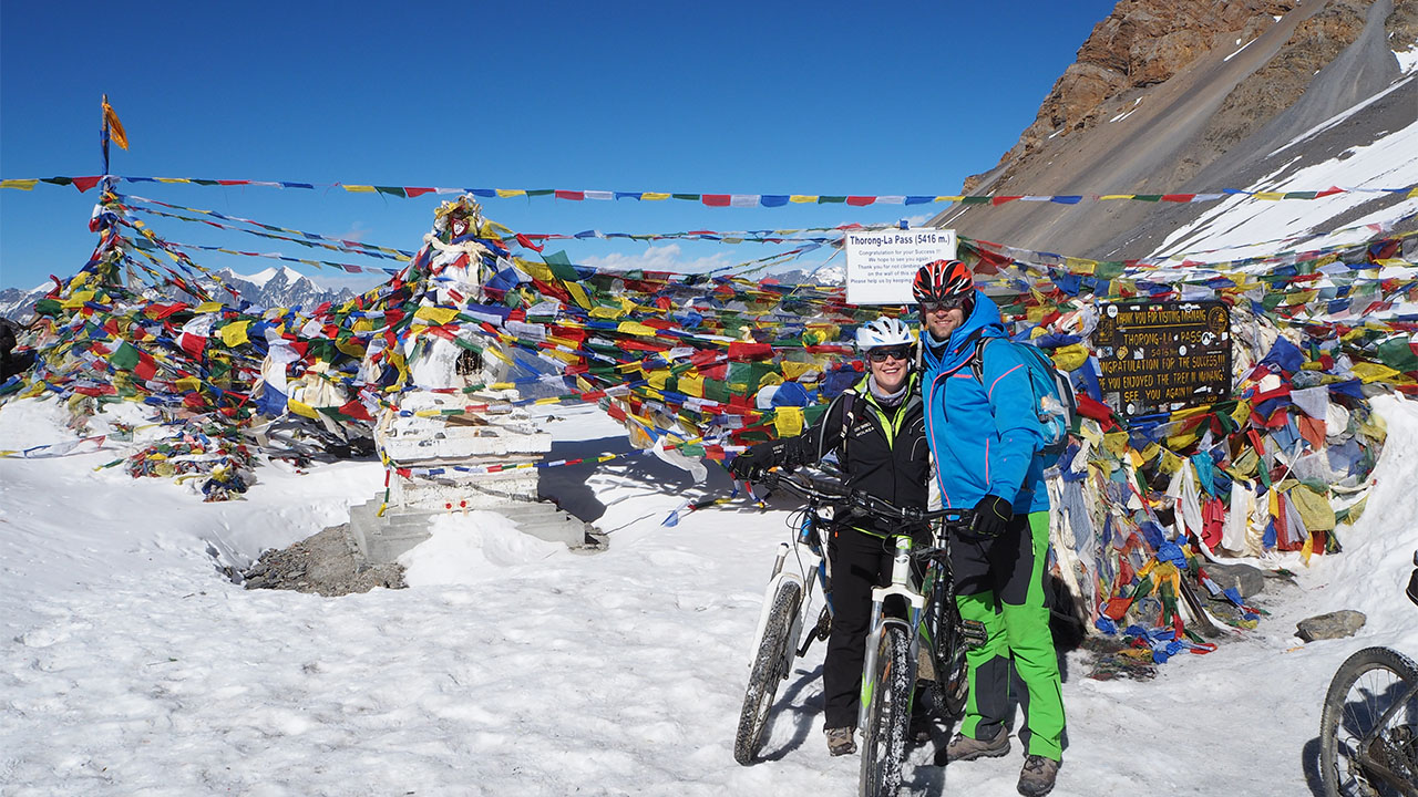 a happy mountain biking couple taking picture with their bikes on the top of Thorong-La, covered with snow and prayer flags