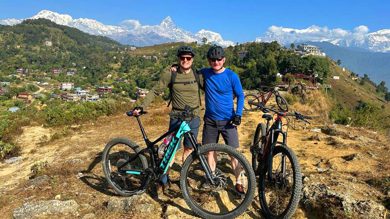 Two mountain bikers are taking a picture with their E-bikes on the side along with the view of Annapurna range and the Fishtail mountain.