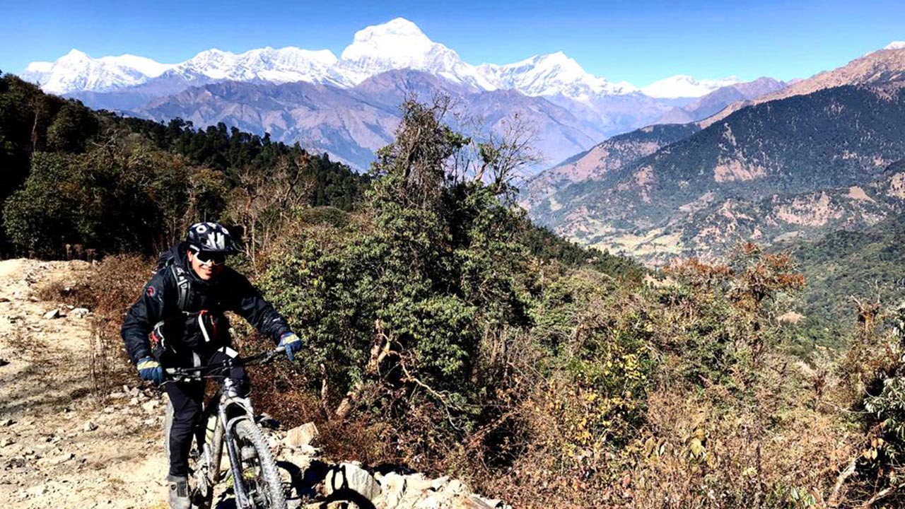 A mountain biking guide in his black outfit is pedaling to Ghorepani and the view of Dhaulagiri mountain range is on the background.