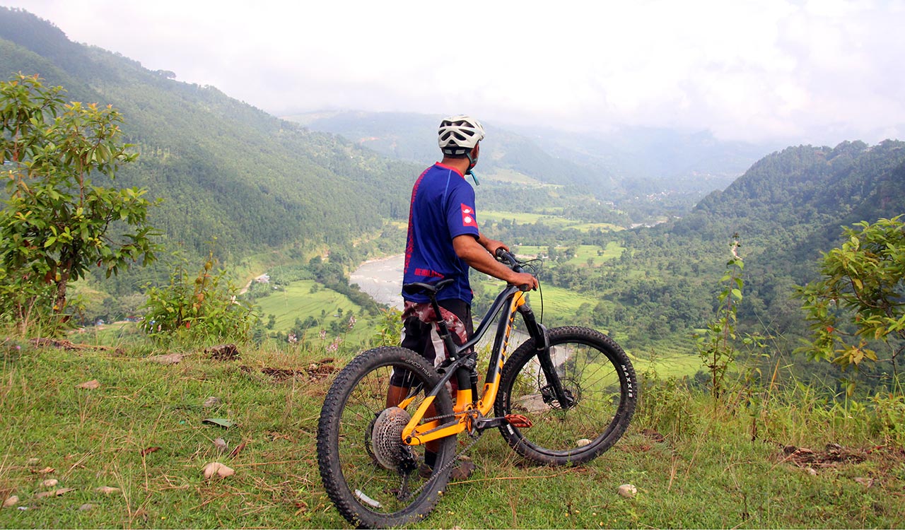 A mountain biker in blue and red T-shirt is observing the view of the Budi Gandaki River valley while holding his bike on his right side.